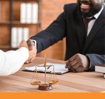 A smiling African-American lawyer shakes hands with a client across a wood table with the scales of justice nearby.