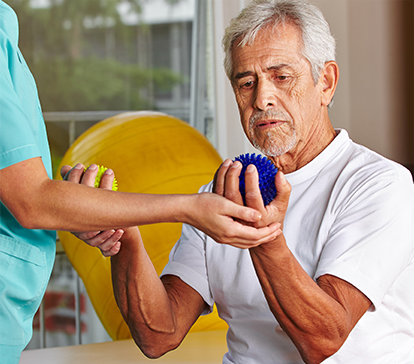 Older male patient in white t-shirt squeezes a blue ball in his left hand and a yellow ball in his right hand