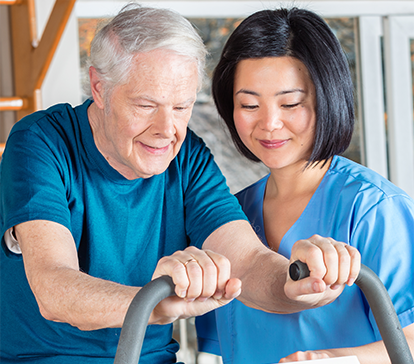 Asian physical therapist works with senior patient to exercise on a stationary bicycle.